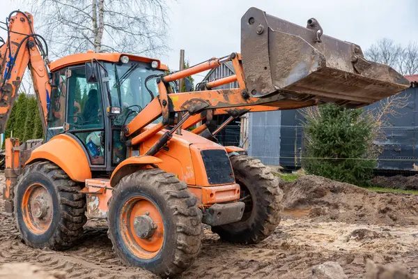 Çamurlu lastikleri ve kaldırılmış kovaları olan turuncu bir kepçe yükleyici inşaat alanında duruyor. Arka planda ağaçlar, çitler ve karanlık bir bina görünüyor..