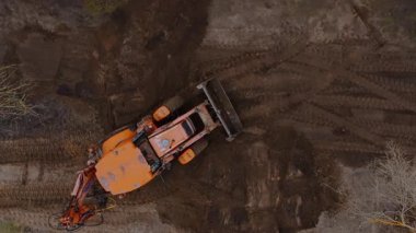 Havadan bakıldığında, çıplak dalları olan seyrek ağaçlarla çevrili, görülebilir lastik izleriyle kaplı toprak bir alanda turuncu bir traktör görülüyor..