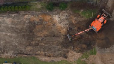 Hava görüntüsü, çimen ve seyrek bitki örtüsüyle çevrili, görünür çit ve malzemelerle çevrili bir inşaat sahasında çalışan turuncu bir kazıcı gösteriyor..