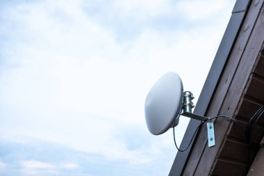 Beyaz dairesel bir Wi-Fi yönlendirici görünür kablolar ve metal bir parantez ile ahşap bir evin dış tarafına monte edilir, parlak, parçalı bulutlu bir gökyüzüne karşı kurulur..