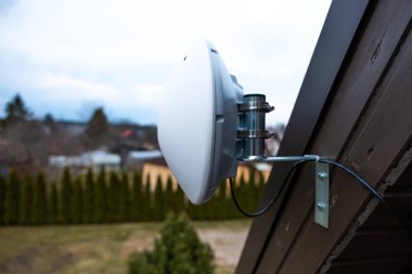 Beyaz kubbe şekilli WiFi yönlendirici ev duvarına metal ayraçlarla monte edilmiş, her zaman yeşil ağaçlara, yerleşim yapılarına ve bulutlu gökyüzüne kurulmuş..