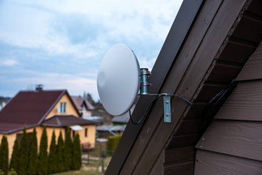 Beyaz dairesel Wi-Fi anteni, eğimli ahşap evin dış tarafına monte edilmiş. Komşu evler, eğimli çatılar ve bulutlu gökyüzünün altında görünen uzun ağaçlar..