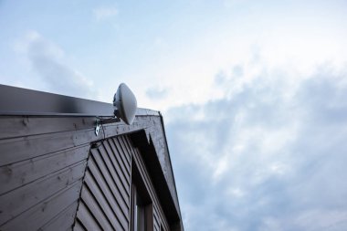 Çatı eğimli, tahta bir evin köşesinde kısmen bulutlu bir gökyüzüne karşı metal bir deste üzerinde beyaz kubbe şekilli Wi-Fi yönlendirici var..