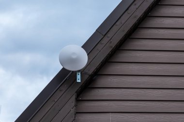 Modern beyaz Wi-Fi yönlendirici, kısmen bulutlu bir gökyüzüne karşı kurulmuş, sessiz kahverengi panelli bir evin eğimli ahşap kaplamasına monte edilmiş..