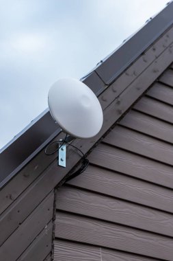 Dairesel beyaz bir Wi-Fi yönlendirici özel bir evin çatı çizgisi yakınındaki eğimli ahşap kaplamalara monte edilir. Gökyüzü hafif bulutlarla kaplıdır..