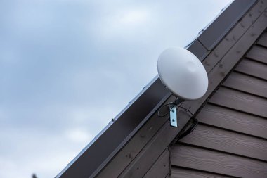 Dairesel beyaz bir Wi-Fi yönlendirici görünür kablolarla eğimli bir çatıya monte edilir. Ev ahşap panellerden oluşuyor ve gökyüzü bulutlu..