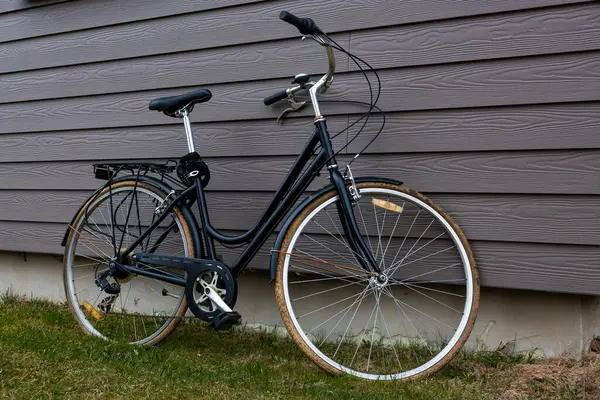 Çerçeveden geçen siyah bir bisiklet gri ahşap bir ev duvarına yaslanır. Üste çim ve toprak görünüyor. Bu da yerleşim yeri olduğunu gösteriyor..