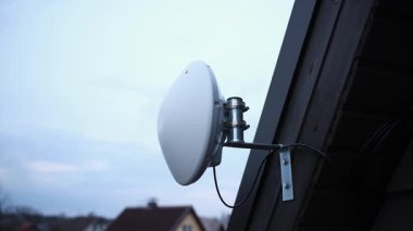 Beyaz dairesel bir Wi-Fi yönlendirici görünür kablolar ve metal bir parantez ile ahşap bir evin dış tarafına monte edilir, parlak, parçalı bulutlu bir gökyüzüne karşı kurulur..
