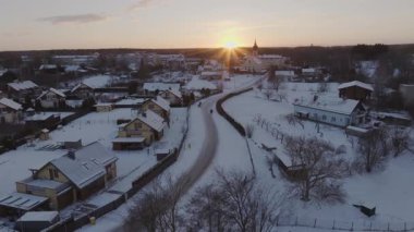 Gün batımında bir kış köyünde dolambaçlı bir yol, yüksek bir kilise, eğimli çatı evleri, çıplak ağaçlar ve altın ışıkta yürüyen insanlar bulunur..