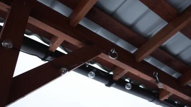 Metal çatılı ve sicim lambalı ahşap bir teras, karla kaplı çatılar, ağaçlar ve kışın kırmızı tuğlalarla kaplı bir bina..