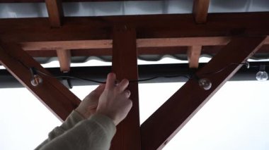 Ellerinizi kapatın ve ahşap bir terasa LED sicim ampulleri takın. Üçgen kirişler ve bulutlu bir gökyüzünün altında eğimli metal bir çatı..