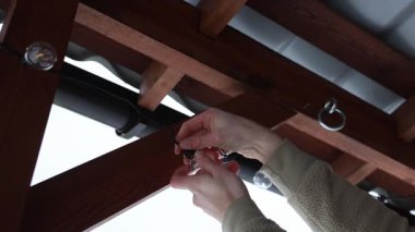 Bej ceketli şahıs, LED ampulleri metal çatılı bir teras pergola üzerinde ayarlıyor. Arka planda karla kaplı çatılar ve çitler görülüyor..