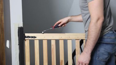 Bir birey, merdivenin tepesinde siyah metal menteşeli ahşap bir güvenlik kapısını güvence altına almak için alet kullanıyor. Duvarda bir ışık düğmesi var..