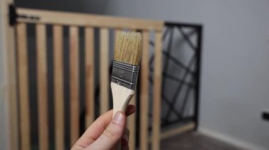 Bir el 5 santimlik doğal bir fırça tutar. Dikey kirişli ve siyah geometrik metal çerçeveli ahşap bir kapı iç mekanda görülebilir..