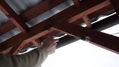 Ellerinizi kapatın ve ahşap bir terasa LED sicim ampulleri takın. Üçgen kirişler ve bulutlu bir gökyüzünün altında eğimli metal bir çatı..