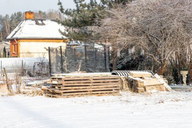 Kar kaplı bir avluda ahşap paletler, beton bloklar ve malzemeler yer alır. Çatı ve bacası eğimli bir ev ağaçların ve yemyeşil ağaçların arasında duruyor..