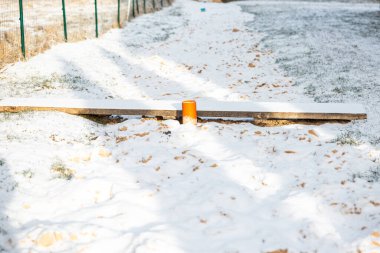 Turuncu destekli ahşap bir tahta, kar tozlu bir alanda tahterevalli oluşturur. Çimenler ve yapraklar görünür, sol tarafında metal bir çit var..