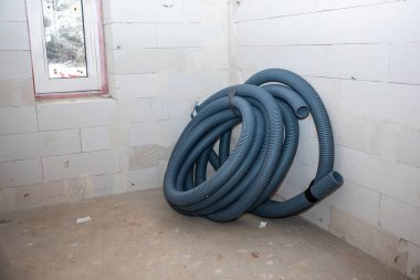 Tamamlanmamış beyaz blok duvarları olan bir oda, sarmalanmış mavi bir tüp, ağaçları gösteren kırmızı çerçeveli bir pencere, ve dağınık enkazlarla dolu çıplak bir zemin..