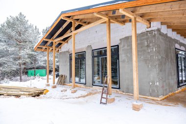 Ahşap kirişleri, beton duvarları ve büyük cam pencereleri olan inşaat halinde bir bina. Arka planda karla kaplı ağaçlar ve yeşil bir konteyner var..