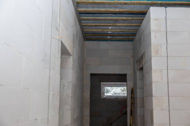 Beyaz beton duvarları, küçük bir penceresi, korunmasız ahşap kirişleri, yalıtımı ve arka planda merdiveni olan bitmemiş bir binanın içi..