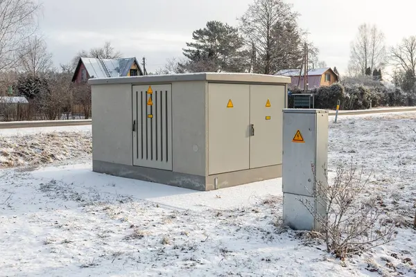 Karla kaplı bir alanda sarı uyarı işaretleri, bir kontrol paneli, seyrek bitki örtüsü ve arka planda eğimli çatıları olan evler bulunan bir hizmet kutusu yer almaktadır..