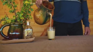 Bir insan kırsal bir testiden bardağa kremalı bir içecek döker. Seramik sürahide bir bitki ve tohumlu bir şişe tahta masada oturur..