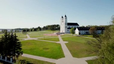 İki kulesi olan büyük beyaz bir kilise, çimenler ve patikalarla çevrili mavi bir gökyüzüne karşı duruyor. İnsanlar uzaktaki ağaçlar ve suyla yakınlarda yürür..