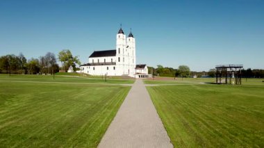 Kırmızı asfalt ve ağaçlarla çevrili, iki kuleli büyük beyaz barok tarzı bir kilise su kenarında duruyor. Birkaç kişi açık mavi gökyüzünün altında yürür..