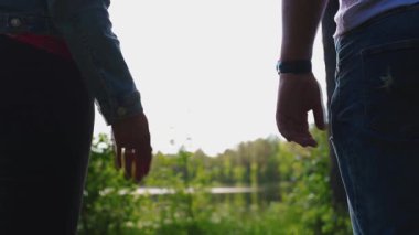 İki birey sırtı kameraya dönük, ağaçlarla çevrili bir su kütlesinin yanında el ele tutuşmuş, güneş ışığı yaprakların arasından süzülüyor..