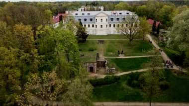 Paslı metal çatılı büyük tarihi bir bina olgun ağaçların ve patikaların arasında duruyor. Ormanlık alanlar ve uzak yapılarla çevrili..