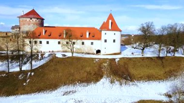 Letonya 'daki Bauska Kalesi' nde kırmızı kiremitli çatılar ve merkezi bir kule yer alıyor. Çıplak ağaçlar ve kar parçaları, açık mavi bir arkaplan ile kışı gösteriyor..