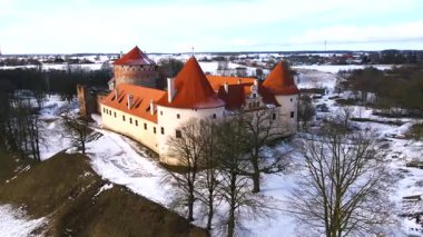 Letonya 'daki Bauska Kalesi' nde çıplak ağaçlar ve açık arazilerle çevrili karlı bir arazide kırmızı kiremitli çatılar ve beyaz taş duvarlar yer alıyor..