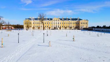 Rundale Sarayı 'nın Barok mimarisi, açık mavi gökyüzünün altında sarı cephesi, geniş pencereleri ve kendine özgü çatısı olan karlı bir manzaraya karşı durmaktadır..