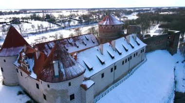Bauska Kalesi Letonya 'da bir tepenin üzerinde duruyor, kırmızı kiremitli çatılar ve taş duvarlar sergileniyor. Kış manzarası, çıplak ağaçlar ve donmuş bir nehirle çevrili..