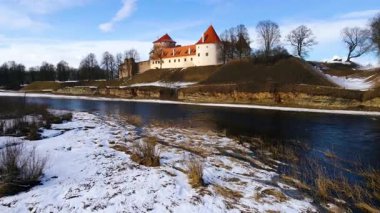 Bauska Kalesi Letonya 'da kırmızı kiremitli çatıları ve beyaz duvarları olan bir tepede duruyor. Karlar toprağı kaplıyor ve çıplak ağaçlar Musa ve Memele nehirlerini kaplıyor..