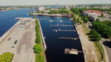 Limanları ve tekneleri sakin sularda, modern bir bina ve kırmızı çatılı tarihi yapılarla çevrili bir yat limanı. Endüstriyel elementler arka planda görünür.