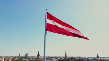 Letonya bayrağı açık bir gökyüzüne karşı dalgalanıyor, Riga 'nın silueti kule ve arka planda tarihi mimariyle birlikte..