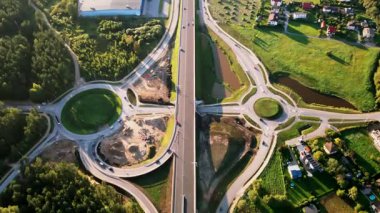 Yükseltilmiş bir otoyol iki kavşakla kesişiyor. Ormanlık alanlar, açık alanlar ve küçük bir su kütlesi, sergilenen altyapı ve doğa ile çevrili..
