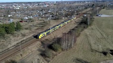 Uzun bir tren, seyrek bitki örtüsü ve arka planda küçük bir yerleşim olan, çıplak ağaçlar ve kahverengimsi otlarla açık alanlarda seyahat eder..