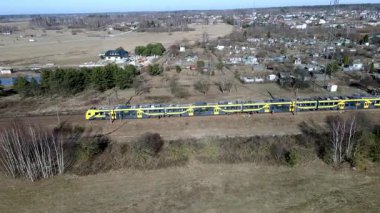 Sarı ve mavi bir tren, dağınık ağaçlar, küçük bir yerleşim yeri ve arka planda büyük bir iletişim kulesi ile açık alanlarda seyahat eder..