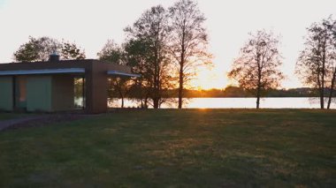 Düz çatılı modern bir bina uzun, yapraksız ağaçların yanında duruyor. Batan güneş, çimenli ön planla çelişen bir su kütlesinin üzerine ılık bir parıltı saçar..
