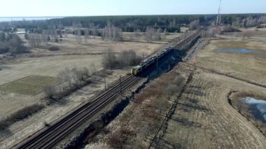 Uzun sarı ve mavi bir tren açık alanlarda ve seyrek bitki örtüsünde seyahat eder. Küçük bir yerleşim yeri solda görünüyor, uzakta ormanlık bir alan var..