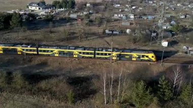Sarı ve siyah bir tren, seyrek bitki örtüsü ve küçük yapılarla kırsal bir alanda demiryolu boyunca hareket eder. Arka planda belirgin bir metal kule var..