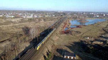 Sarı bir tren, küçük su ve kırmızı bitki örtüsüyle çevrili kırsal bir alanda demiryolu boyunca seyahat eder. Açık mavi gökyüzünün altında küçük bir kasaba görülebilir..