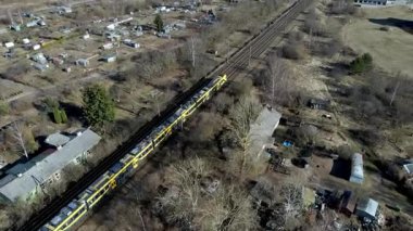Sarı bir tren, sol ve sağ tarafındaki kırmızı ve gri çatıları olan modern binalarla çevrili kırsal bir alanda ilerliyor..