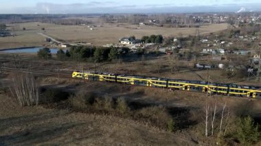 Sarı ve siyah bir tren açık araziler ve dağınık ağaçlar arasında hareket eder. Arka planda evleri ve yüksek bir iletişim kulesi olan küçük bir yerleşim yeri görülüyor..