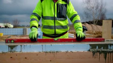 Yeşil, yüksek görüş mesafesi olan bir inşaat işçisi duvarda kırmızı bir ruh seviyesi kullanır. Sitede toprak yığınları ve aktif çalışmayı gösteren bulutlu bir gökyüzü yer alıyor..