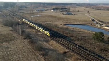 Modern bir sarı ve siyah tren seyrek ağaçlar ve küçük bir gölet ile düz araziler boyunca seyahat eder, uzak binalar ve bir ağaçlık alana karşı kurulur..