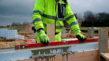 Sarı ve gri ceketli bir inşaat işçisi beton bir yüzey üzerinde kırmızı ruh seviyesi kullanır. Kum yığınları ve malzemeler bulutlu bir gökyüzünün altında görünür..