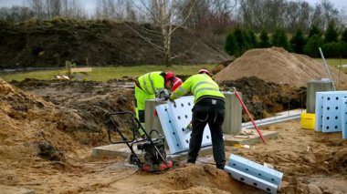 Yüksek görüş teçhizatı ve kırmızı kasklı iki işçi büyük paneller kuruyor. Kum toprağı, toprak yığınları ve inşaat ekipmanları görülebilir, arka planda ağaçlar var..